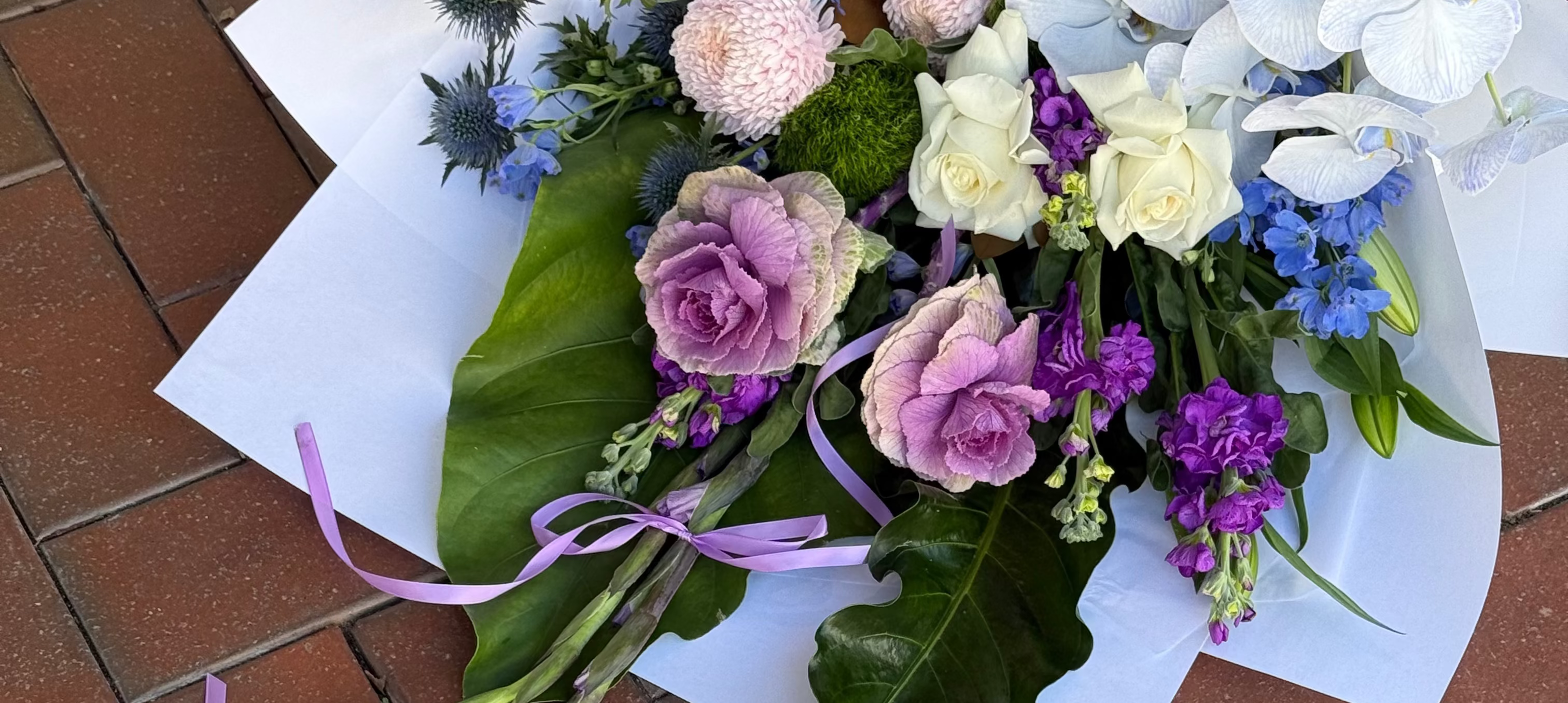 A large, vibrant mixed floral bouquet featuring purple roses, white roses, blue flowers, and green leaves, wrapped in white paper with a purple ribbon, laid flat on a red brick surface.